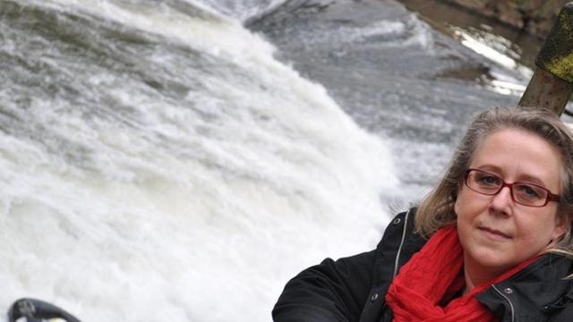 Das Bild zeigt die Krimi-Bestsellerautorin Elke Pistor am "Tatort" ihrer Kindheit: eine Flusswehr in Gemünd in der Eifel. Genau hier spielt einer ihrer Romane, und hier liegt auch der Kriminal-Lehrpfad.
