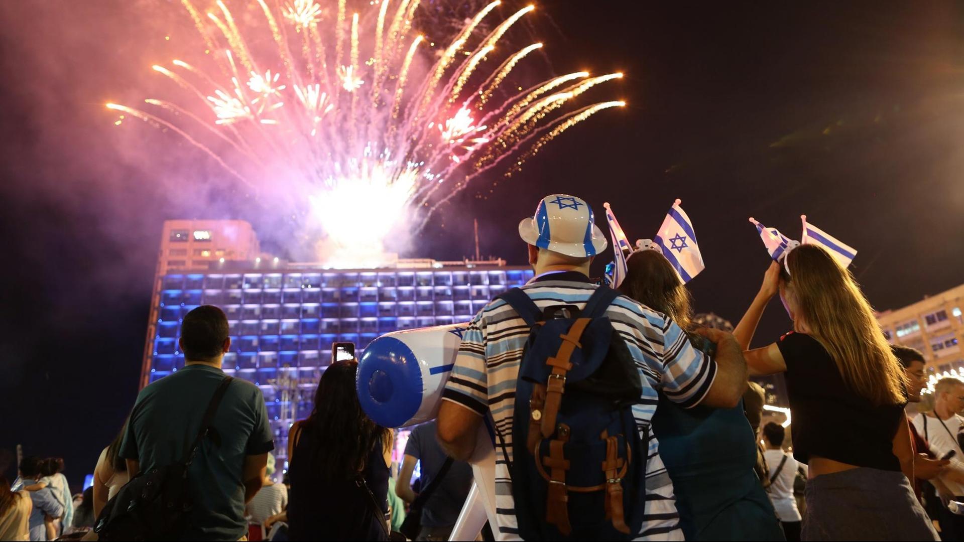 Das Bild zeigt Menschen auf dem Rabin-Platz in Tel Aviv. Sie schauen ein Feuerwerk an, einer trägt einen Hut in den Farben der Israelflagge. Das Bild zeigt Menschen auf dem Rabin-Platz in Tel Aviv. Sie schauen ein Feuerwerk an, einer trägt einen Hut in den Farben der Israelflagge.