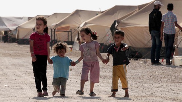 Vier Kinder, 2 größere Mädchen und 2 kleinere Jungs, gehen Hand in Hand auf einem Weg im Flüchtlingslager, im Hintergrund sind Zelte zu sehen