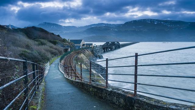 Eine Brücke in Wales führt über einen Fluss entlang der Küste. Im Hintergrund sind Berge zu sehen.