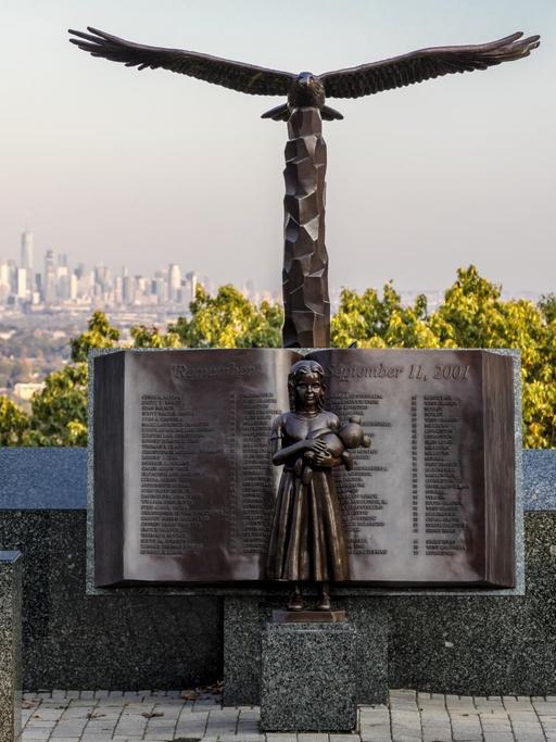 Bronzefigur eines aufgeschlagenen Buches mit den Namen der Opfer von 9/11. Vor dem Buch die Figur eines Mädchens, dass einen Teddy hält. Darüber die Figur eines Adlers. Im Hintergrund ist die Silhouette von New York zu sehen. Eagle Rock Reservation, New Jersey, USA.