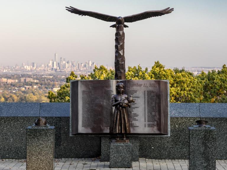 Bronzefigur eines aufgeschlagenen Buches mit den Namen der Opfer von 9/11. Vor dem Buch die Figur eines Mädchens, dass einen Teddy hält. Darüber die Figur eines Adlers. Im Hintergrund ist die Silhouette von New York zu sehen. Eagle Rock Reservation, New Jersey, USA.