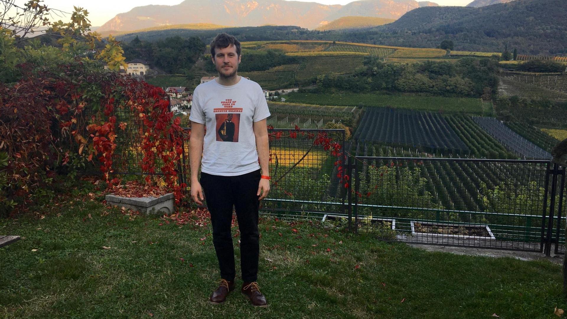 Auf dem Bild ist der Musiker Marcus Hamblett zu sehen. Er steht vor einer Berglandschaft und lächelt in die Kamera Auf dem Bild ist der Musiker Marcus Hamblett zu sehen. Er steht vor einer Berglandschaft und lächelt in die Kamera