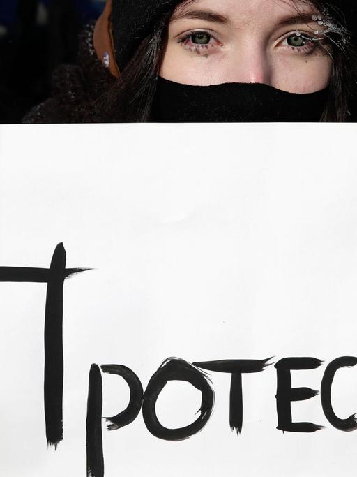 Eine junge Frau mit Wollhandschuhen, Mütze und Mundschutz hält ein Schilf, auf dem in kyrillischer Schrift "Protest" steht.