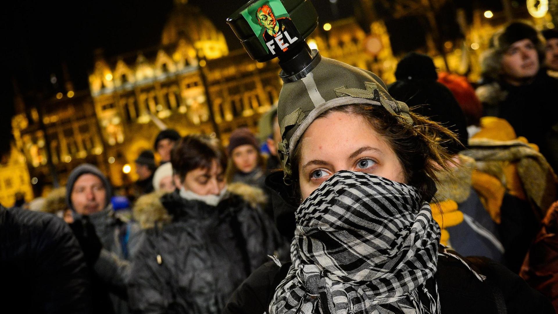 Eine Demonstrantin ist mit einem Tuch vermummt vor dem ungarischen Parlament in Budapest. Um sie herum stehen weitere Demonstranten.