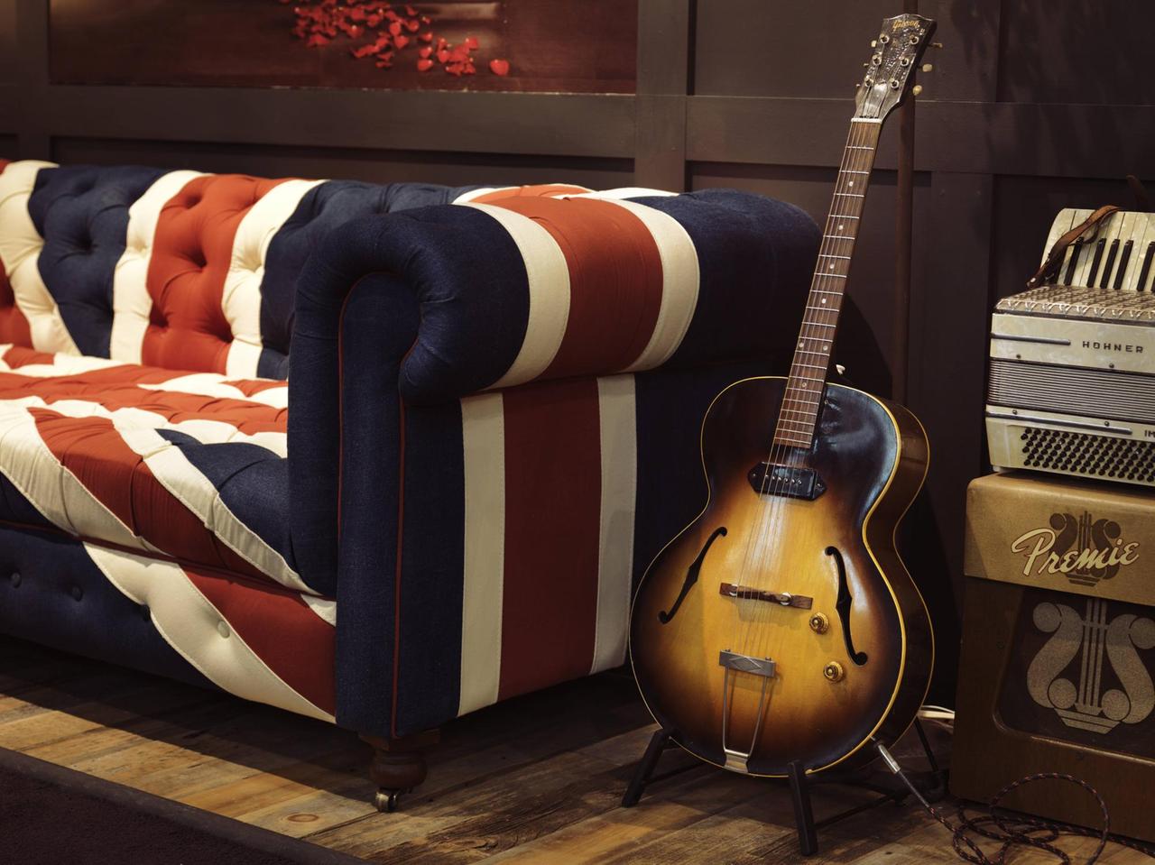 Eine alte akustische Gibson-Gitarre steht auf einem Ständer neben einem Sofa mit dem Union Jack als Muster.