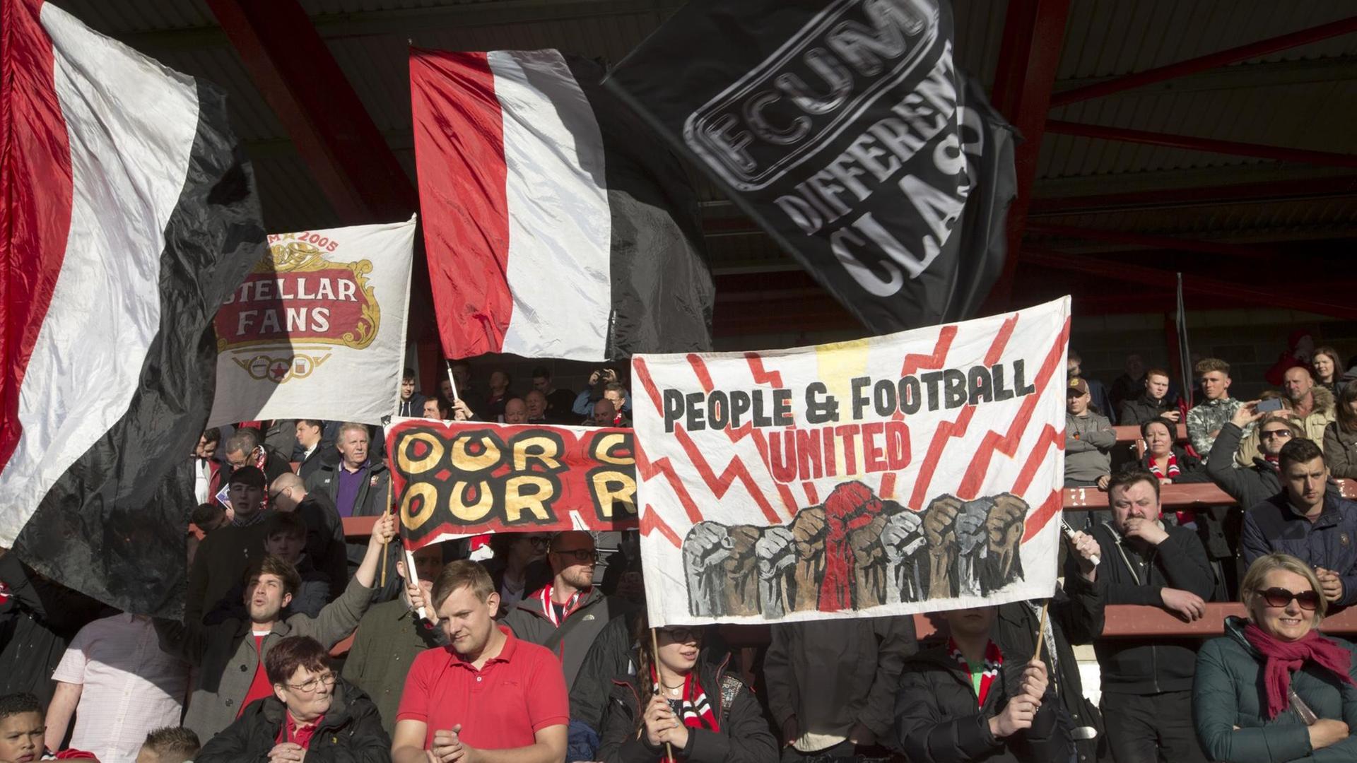 Fans von FC United of Manchester schwenken Banner im Broadhurst Park Stadion. Fans von FC United of Manchester schwenken Banner im Broadhurst Park Stadion.