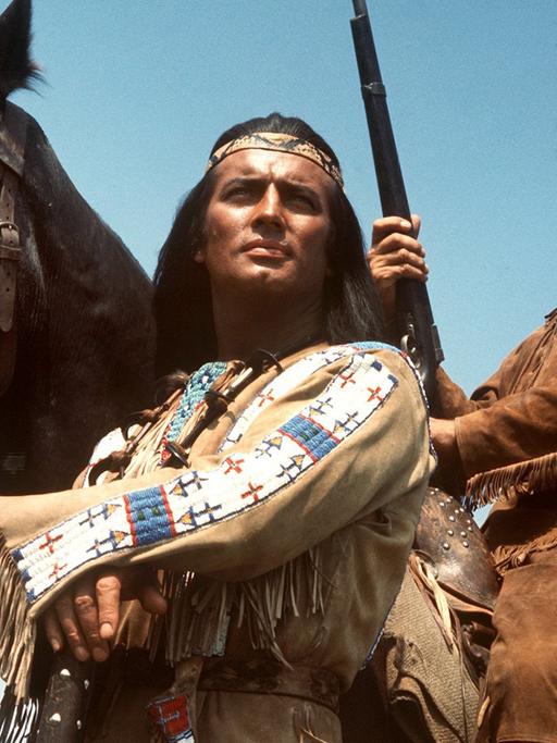 Pierre Brice (l) als Apachen-Häuptling Winnetou und Lex Barker als sein Blutsbruder Old Shatterhand in einer Szene des Karl-May-Films "Im Tal des Todes". Lex Barker sitzt auf einem Pferd mit einem Gewehr in der Hand. Beide schauen in die Ferne.