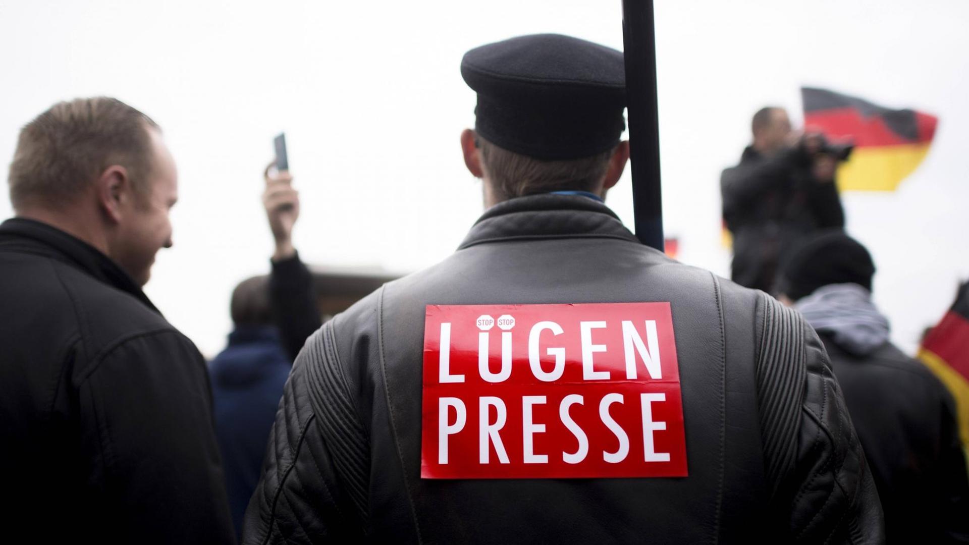 Ein Mann ist von hinten zu sehen. Er trägt eine Schiebermütze und auf seinem Rücken steht "Lügenpresse". Dahinter ist eine Deutschlandfahne zu sehen.