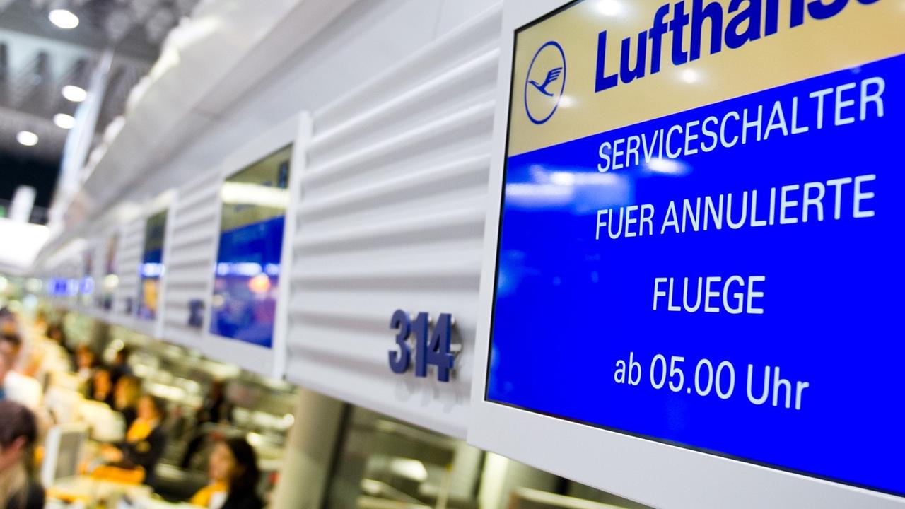 An einem Flugschalter der Lufthansa in Frankfurt am Main ist auf dem Display zu lesen: "Serviceschalter für annulierte Flüge ab 5 Uhr." Vor dem Schalter sind unscharf Fluggäste zu erkennen; dahinter sitzen zwei Service-Angestellte der Lufthansa. An einem Flugschalter der Lufthansa in Frankfurt am Main ist auf dem Display zu lesen: "Serviceschalter für annulierte Flüge ab 5 Uhr." Vor dem Schalter sind unscharf Fluggäste zu erkennen; dahinter sitzen zwei Service-Angestellte der Lufthansa.