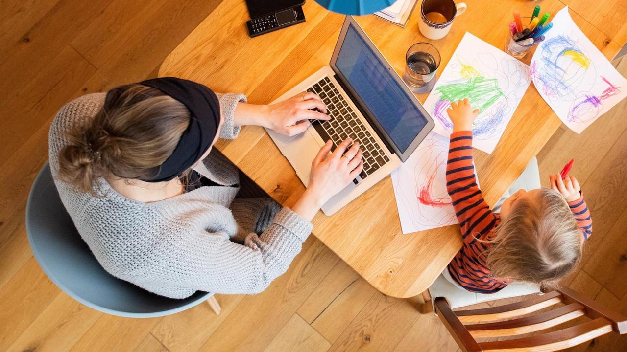 Ein zweijähriges Kind malt ein Bild, während seine Mutter im Homeoffice an einem Laptop arbeitet. Ein zweijähriges Kind malt ein Bild, während seine Mutter im Homeoffice an einem Laptop arbeitet.