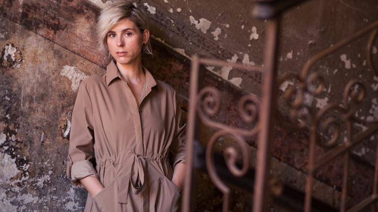Die Musikerin steht in einem hellbraunen Kleid vor einer abblätternden Wand und einem gußeisernen Geländer Die Musikerin steht in einem hellbraunen Kleid vor einer abblätternden Wand und einem gußeisernen Geländer