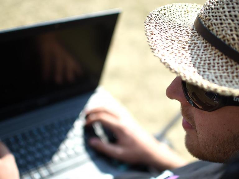 Ein Mann mit Strohhut, Sonnenbrille und Notebook auf dem Schoß