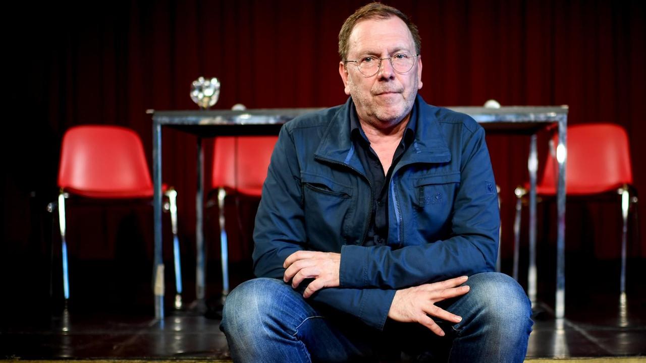 Rene Pollesch, Theaterregisseur und Autor, sitzt am Rande einer Pressekonferenz zur "Zukunft der Volksbühne". Er sitzt auf einer niedrigen Bühne, im Hintergrund sind ein Tisch und vier Stühle mit roten Kunststoff-Sitzen zu sehen. Rene Pollesch, Theaterregisseur und Autor, sitzt am Rande einer Pressekonferenz zur "Zukunft der Volksbühne". Er sitzt auf einer niedrigen Bühne, im Hintergrund sind ein Tisch und vier Stühle mit roten Kunststoff-Sitzen zu sehen.