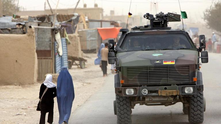 Ein Bundeswehrfahrzeug fährt auf einer Straße in Afghanistan an zwei Frauen vorbei, die eine trägt eine Burka, die andere ein Kopftuch. 