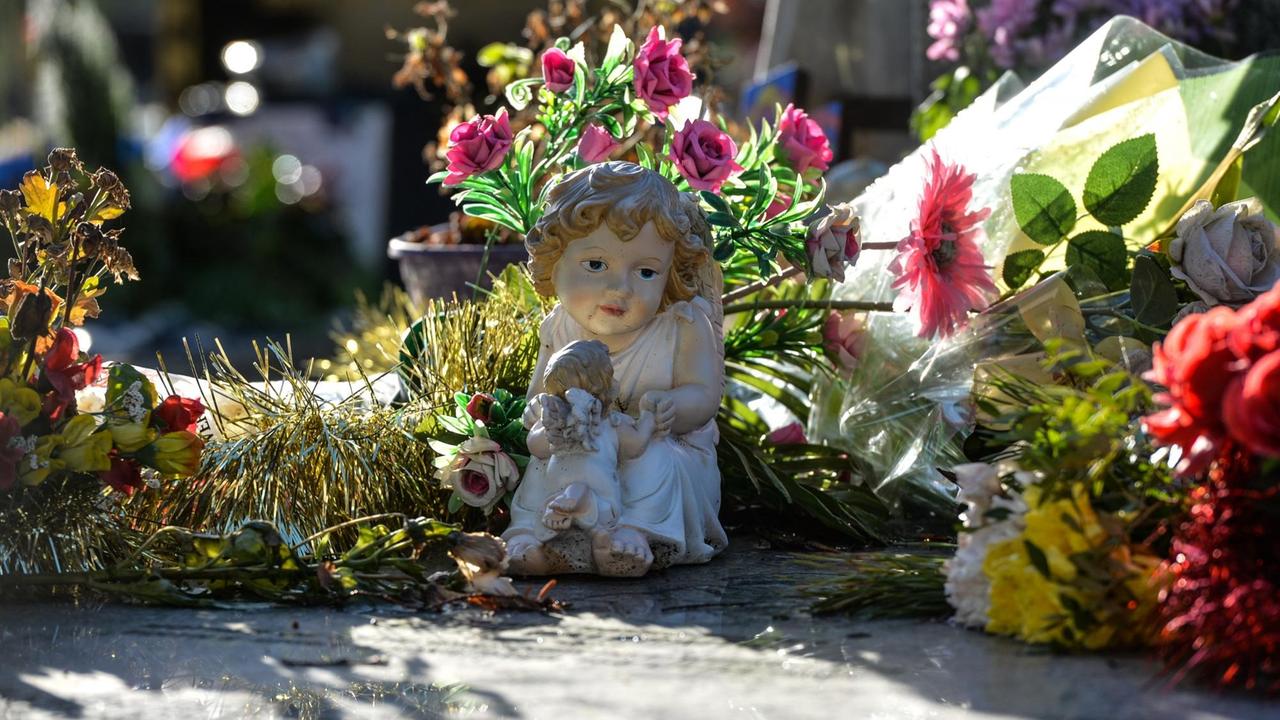 Figuren und Blumen stehen und liegen am Boden, in der Mitte eine Engelsfigur, die ein Kind auf dem Arm hat Figuren und Blumen stehen und liegen am Boden, in der Mitte eine Engelsfigur, die ein Kind auf dem Arm hat