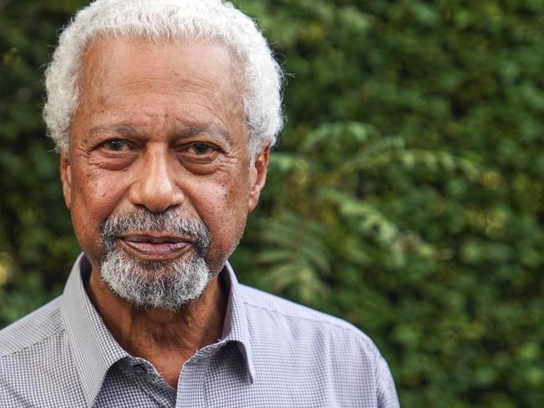 Der Literaturnobelpreisträger Abdulrazak Gurnah blickt in die Kamera. Hinter ihm sind Bäume zu sehen; der Autor trägt ein graues Hemd.