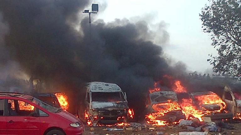 Mehrere Fahrzeuge brennen, schwarzer Qualm zieht in den Himmel, im Vordergrund links ein teilweise ausgebranntes rotes Auto Mehrere Fahrzeuge brennen, schwarzer Qualm zieht in den Himmel, im Vordergrund links ein teilweise ausgebranntes rotes Auto