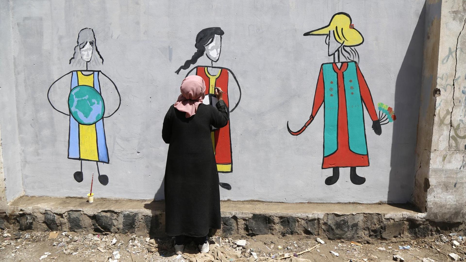 Eine Frau im Jemen malt zum Internationalen Frauentag ein Graffiti dreier Frauenfiguren an eine Wand. Eine Frau im Jemen malt zum Internationalen Frauentag ein Graffiti dreier Frauenfiguren an eine Wand.