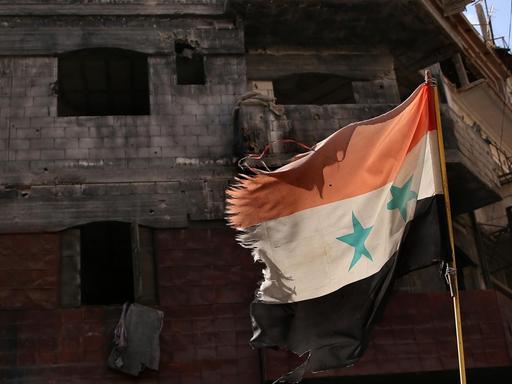 Eine an den Rändern zerfetzte syrische Fahne wehr vor einem zerstörten Gebäude in Homs