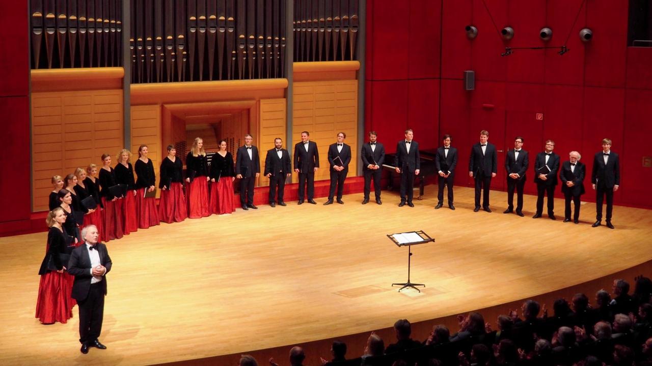 Der Chor steht mit seinem Leiter im Halbkreis auf einer Bühne unter einer modernen Orgel.