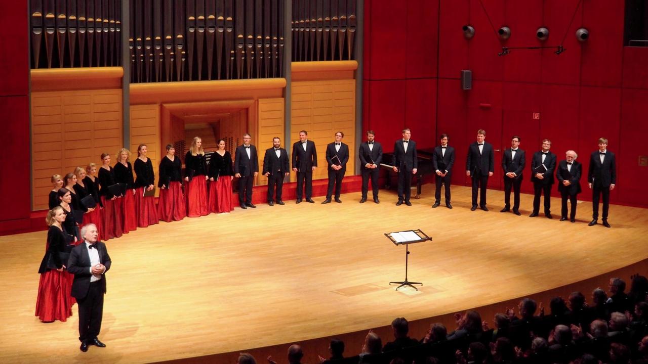 Der Chor steht mit seinem Leiter im Halbkreis auf einer Bühne unter einer modernen Orgel. Der Chor steht mit seinem Leiter im Halbkreis auf einer Bühne unter einer modernen Orgel.