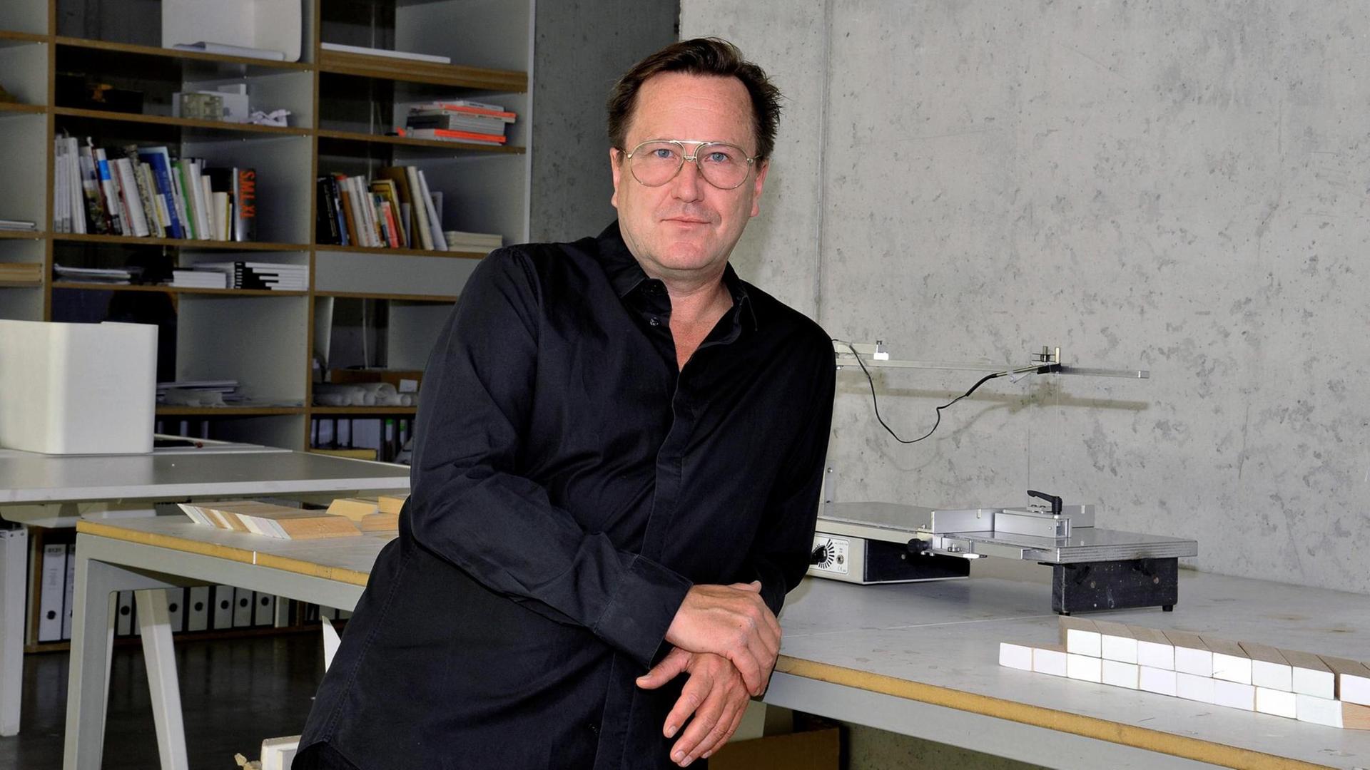 Arno Brandlhuber steht, mit dem Rücken an einen Arbeitstisch gelehnt, in seinem Studio. Im Hintergrund ist eine Sichtbeton-Wand und ein Bücherregal zu sehen.