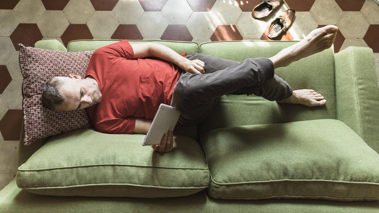 EIn Mann im roten T-Shirt liegt auf einem grünen Sofa und liest auf einem Tablet. EIn Mann im roten T-Shirt liegt auf einem grünen Sofa und liest auf einem Tablet.