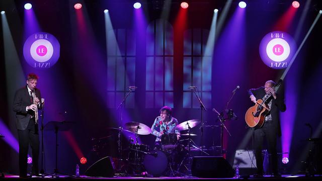 Von links sind auf einer lila eingeleuchteten Konzertbühne ein Tenorsaxofonist, ein Schlagzeuger und ein Gitarrist zu sehen, letzterer spielt auf seinem Instrument mit einem Geigenbogen. Körperhaltungen und Gesichtsausdrücke verraten Egagement und Erregung.
