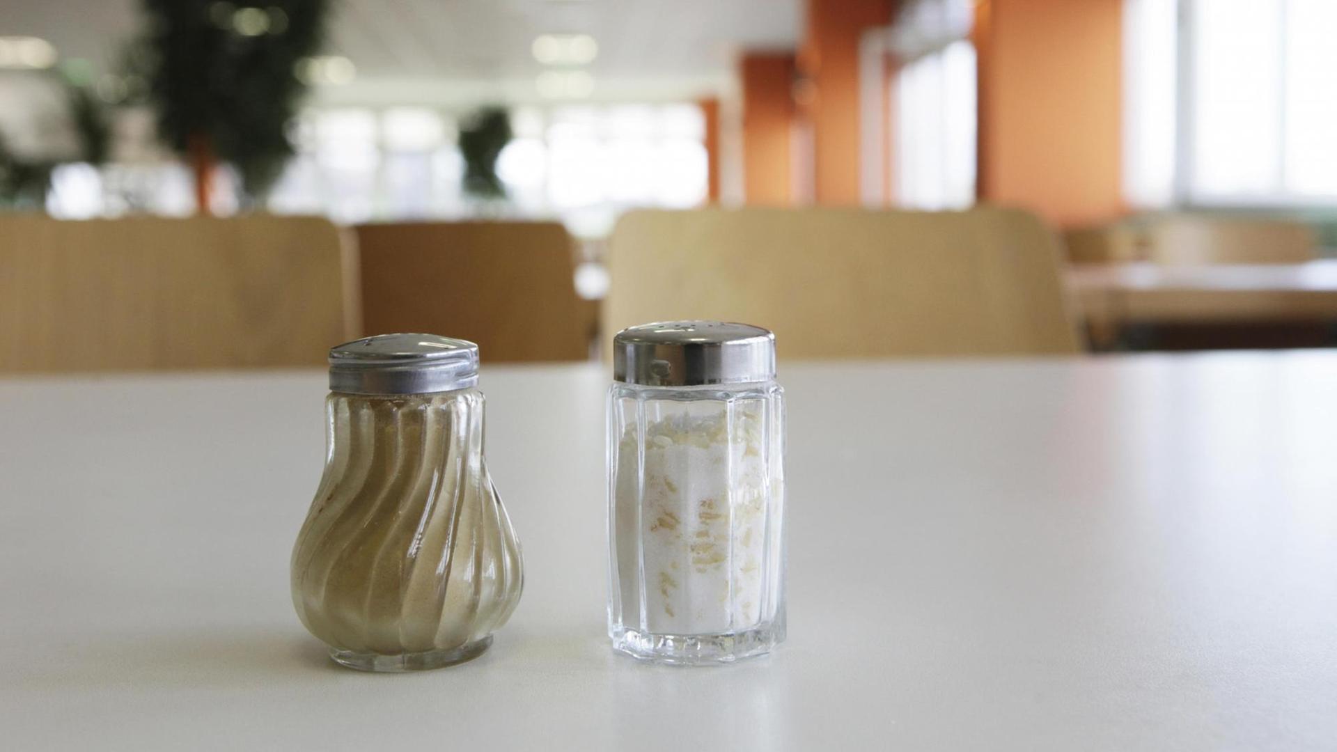 Groß im Bildvordergrund stehen ein Salz- und ein Pfefferstreuer nebeneinander auf dem Esstisch in einer Kantine. Groß im Bildvordergrund stehen ein Salz- und ein Pfefferstreuer nebeneinander auf dem Esstisch in einer Kantine.
