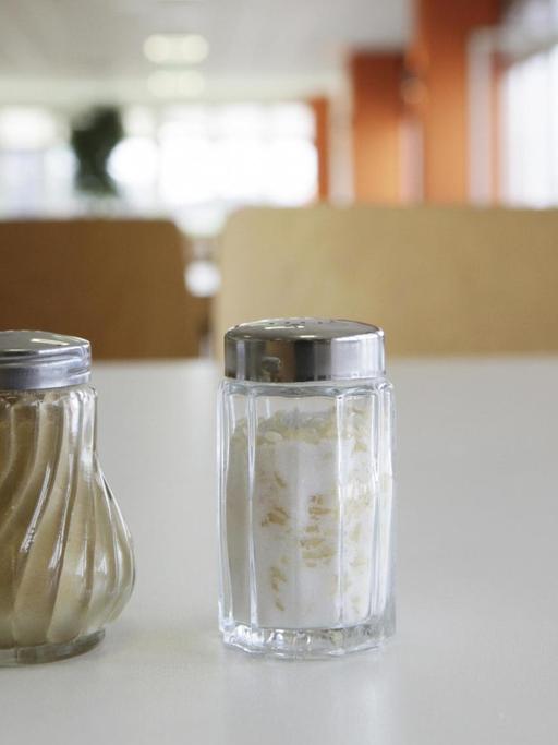 Groß im Bildvordergrund stehen ein Salz- und ein Pfefferstreuer nebeneinander auf dem Esstisch in einer Kantine.