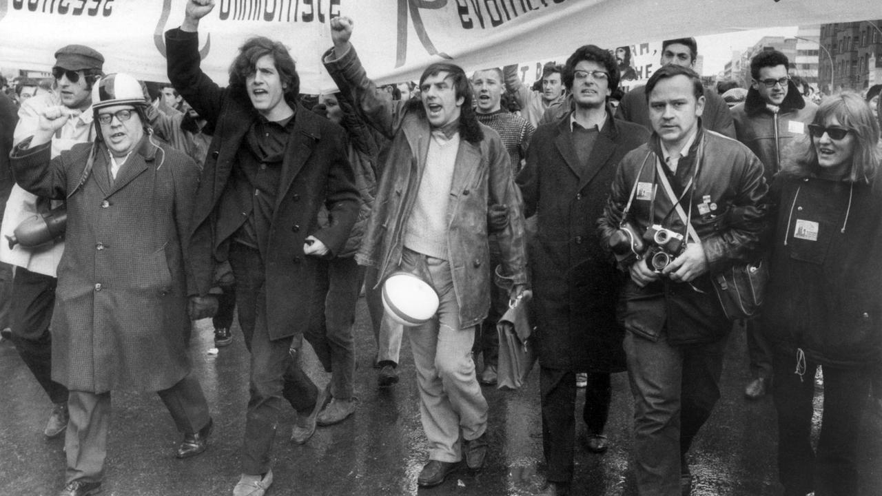Ein Schwarzweiß-Foto zeigt etwa ein Dutzend Demonstrierende unter einem großen Banner schierend und skandierend, die rechte Faust geballt erhoben Ein Schwarzweiß-Foto zeigt etwa ein Dutzend Demonstrierende unter einem großen Banner schierend und skandierend, die rechte Faust geballt erhoben