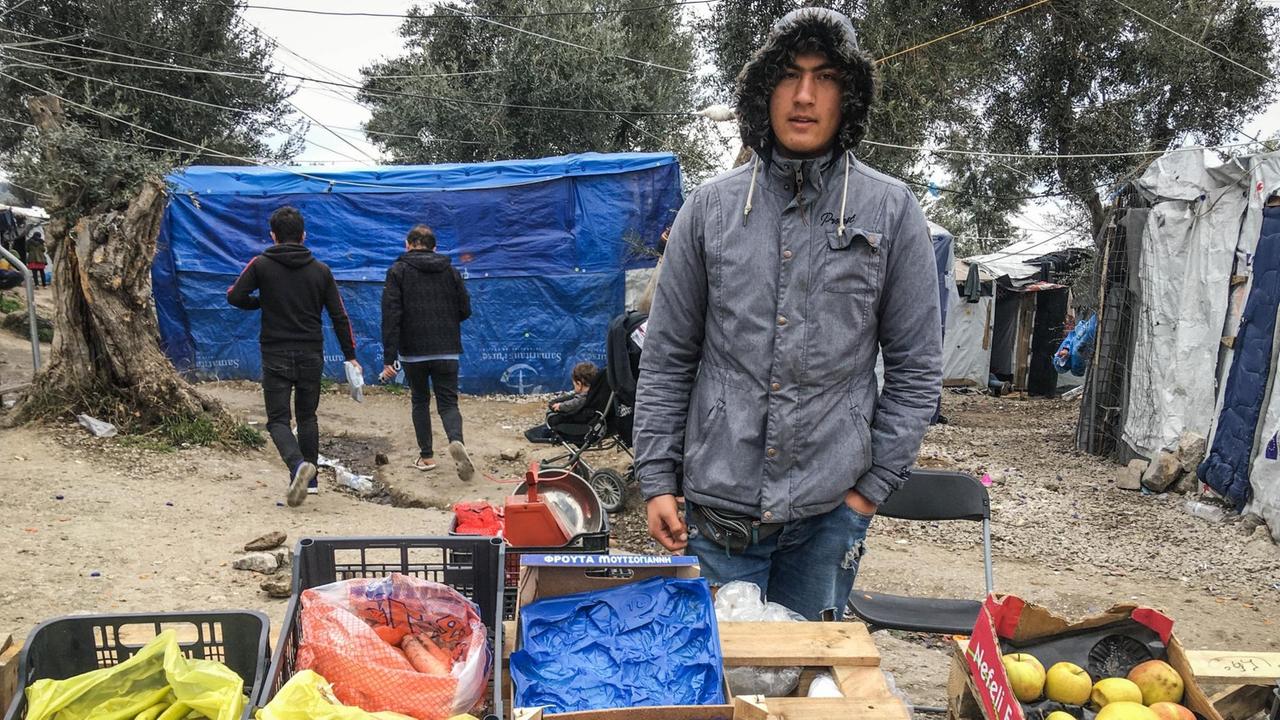 Ein junger Mann in grauem Parka steht vor einem Tisch aus Holzpaletten, auf dem einige Plastikboxen mit Äpfeln, Mandarinen, Karotten und Peperoni zum Verkauf angeboten werden. Ein junger Mann in grauem Parka steht vor einem Tisch aus Holzpaletten, auf dem einige Plastikboxen mit Äpfeln, Mandarinen, Karotten und Peperoni zum Verkauf angeboten werden.
