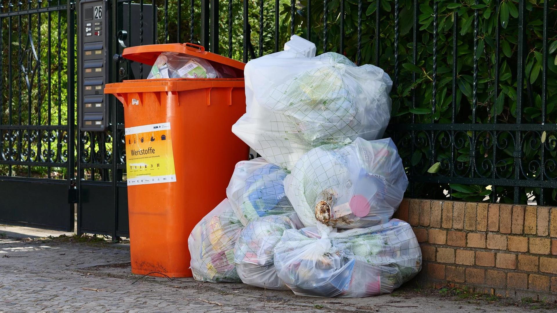 An einem Zaun stehen und liegen Säcke mit Plastikmüll und Verpackungen und es steht eine Tonne voller Wertstoffmüll zur Abholung bereit. An einem Zaun stehen und liegen Säcke mit Plastikmüll und Verpackungen und es steht eine Tonne voller Wertstoffmüll zur Abholung bereit.