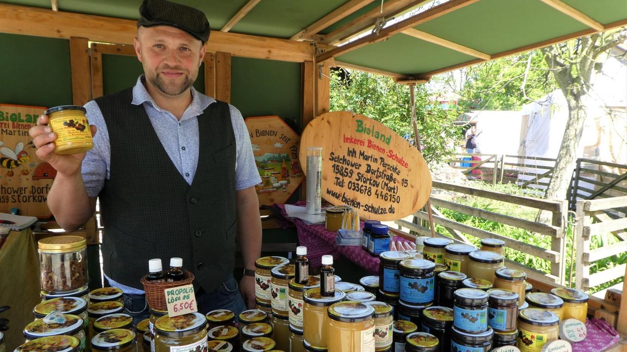 Junger Mann hinter einem Stand mit lauter Bienenprodukten, er hält ein Honigglas in der Hand. Junger Mann hinter einem Stand mit lauter Bienenprodukten, er hält ein Honigglas in der Hand.