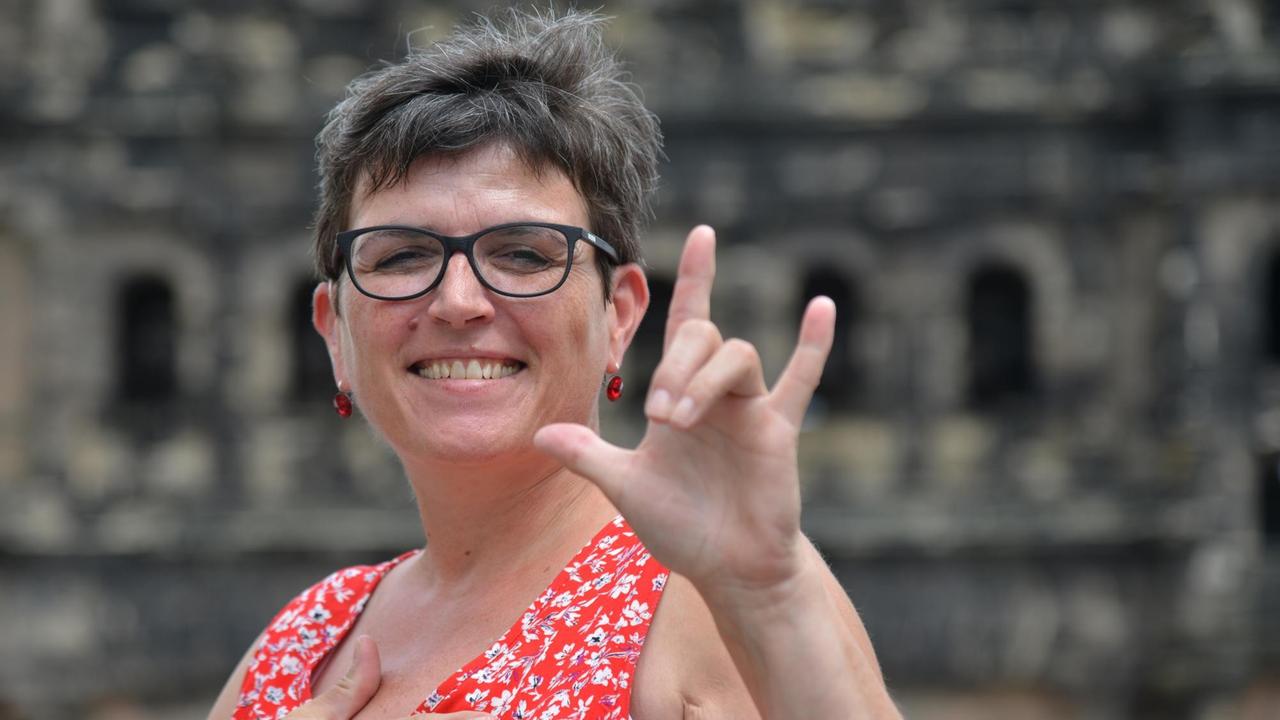 Städteführerin Traudel Theisen zeigt am 31.07.2017 vor der Porta Nigra in Trier per Gebärdensprache das Zeichen für «I love you». Die 50-Jährige ist eine von vier frisch ausgebildeten Städteführerinnen in der Moselstadt, die neuerdings Führungen in Gebärdensprache anbieten. (zu dpa «Trierer Geschichte mit Hand und Fuß: Gehörlose führen Gehörlose» vom 04.08.2017) Foto: Harald Tittel/dpa | Verwendung weltweit Städteführerin Traudel Theisen zeigt am 31.07.2017 vor der Porta Nigra in Trier per Gebärdensprache das Zeichen für «I love you». Die 50-Jährige ist eine von vier frisch ausgebildeten Städteführerinnen in der Moselstadt, die neuerdings Führungen in Gebärdensprache anbieten. (zu dpa «Trierer Geschichte mit Hand und Fuß: Gehörlose führen Gehörlose» vom 04.08.2017) Foto: Harald Tittel/dpa | Verwendung weltweit