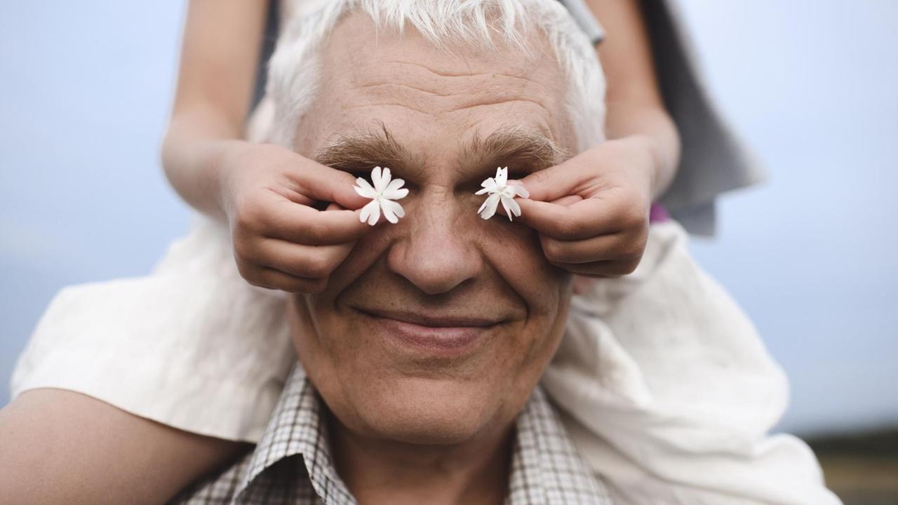 Ein lachender Großvater trägt ein Mädchen auf den Schultern. Es hält ihm zwei weiße Blumen vor die Augen. Ein lachender Großvater trägt ein Mädchen auf den Schultern. Es hält ihm zwei weiße Blumen vor die Augen.