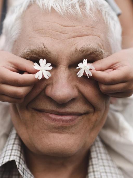 Ein lachender Großvater trägt ein Mädchen auf den Schultern. Es hält ihm zwei weiße Blumen vor die Augen.