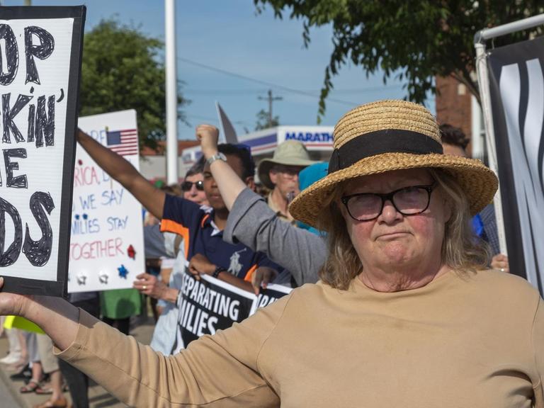 Die ältere Frau mit Strohhut hält das Schild und blickt in die Kamera. Dahinter sieht man weitere Demonstranten mit Schildern am Straßenrand.