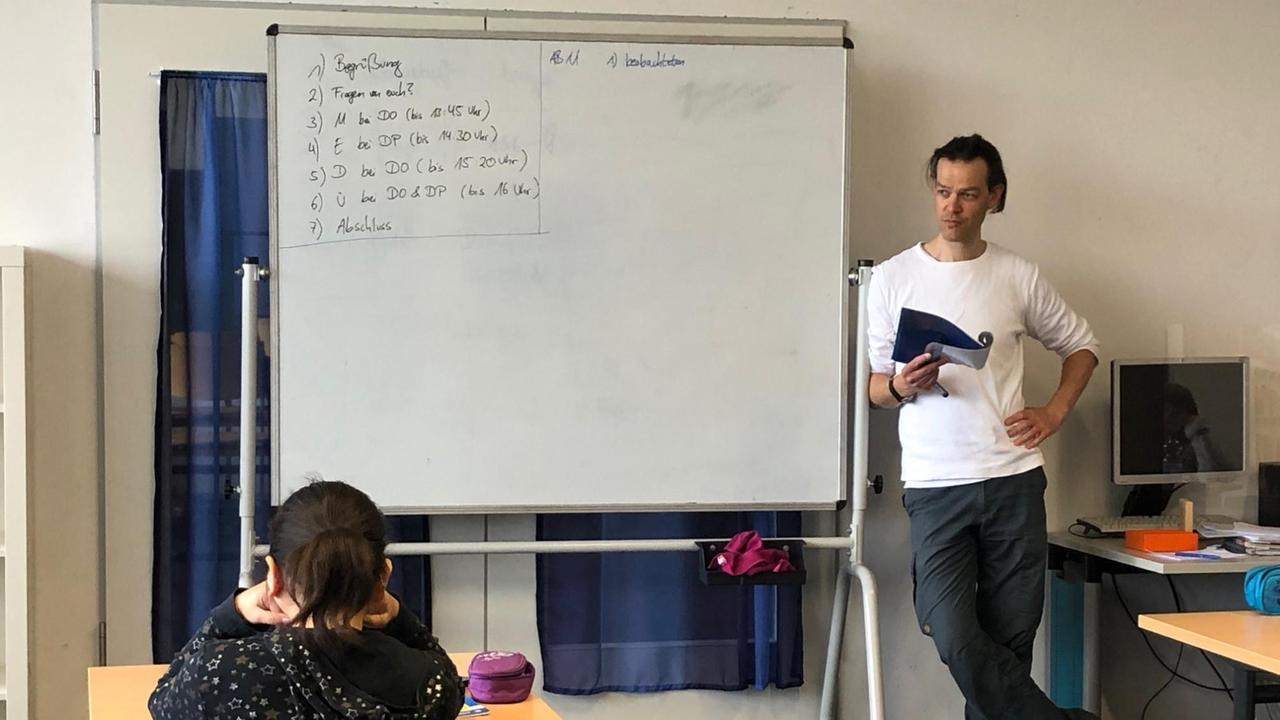 Ein junger Lehrer im weißen T-Shirt und dunkler Hose lehnt in einem Klassenzimmer an der Tafel. Ein junger Lehrer im weißen T-Shirt und dunkler Hose lehnt in einem Klassenzimmer an der Tafel.