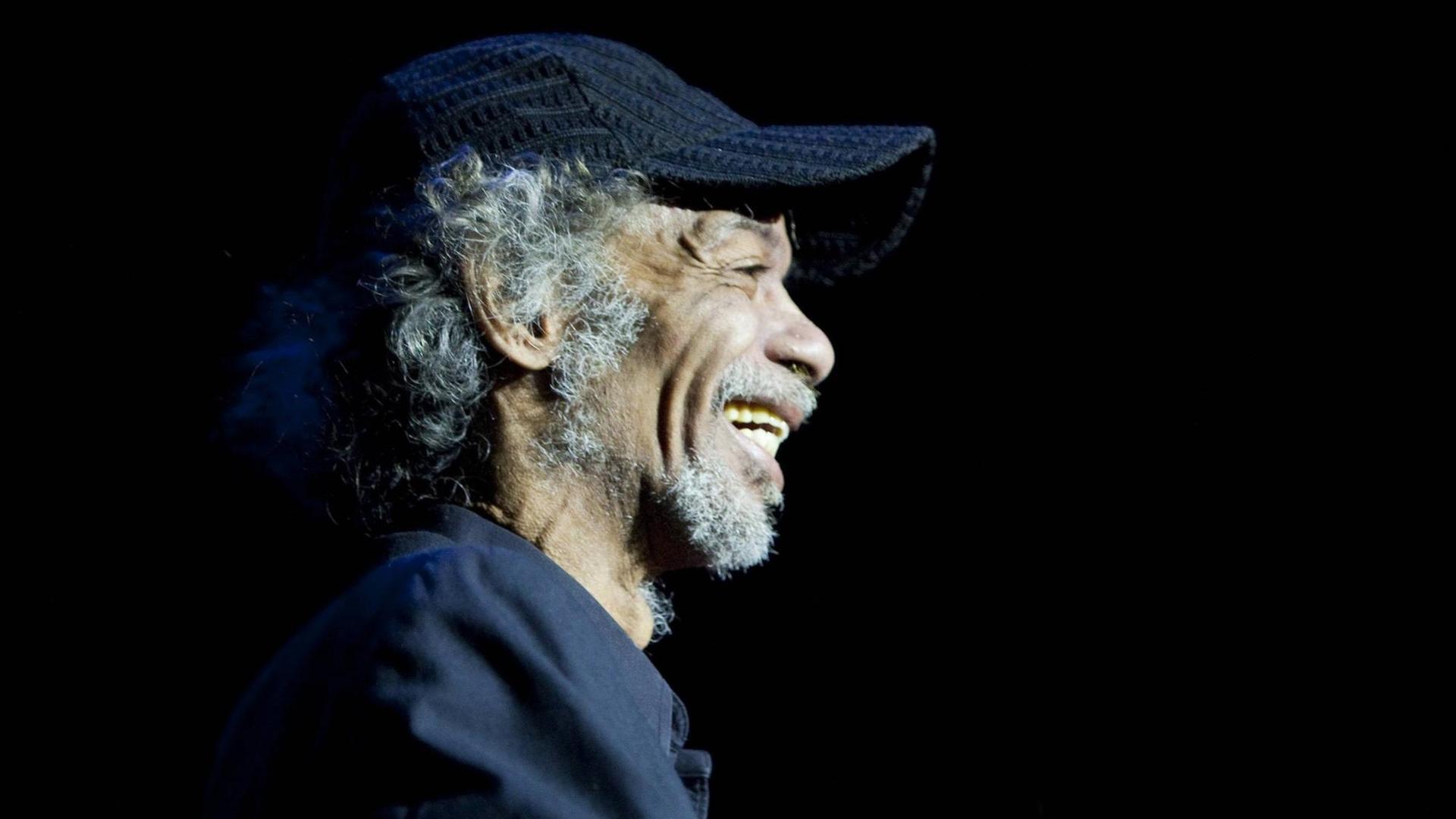 Gil Scott Heron steht lächelnd auf der Bühne und trägt ein Basecape. Gil Scott Heron steht lächelnd auf der Bühne und trägt ein Basecape.