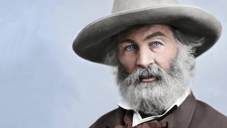 Ein coloriertes Porträt des Dichters Walt Whitman. Er sitzt mit einem Hut mit breiter Krempe auf einen Stuhl und schaut in die Kamera. Seine Hände sind in den Manteltaschen.