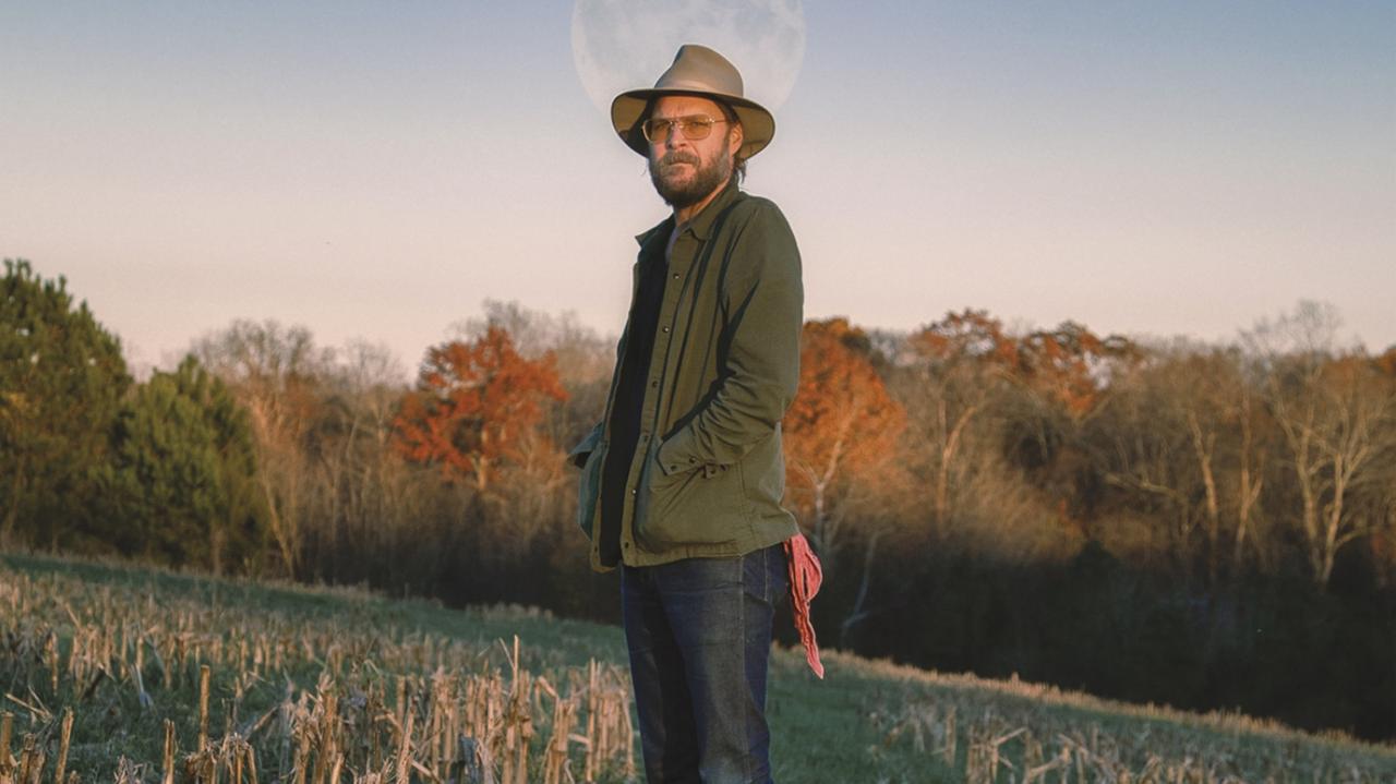 Ein Mann in einer grünen Jacke mit Hut und Brille steht auf einem abgemähten Feld. Im Hintergrund stehen rotgefärbte Bäume.. Ein Mann in einer grünen Jacke mit Hut und Brille steht auf einem abgemähten Feld. Im Hintergrund stehen rotgefärbte Bäume..