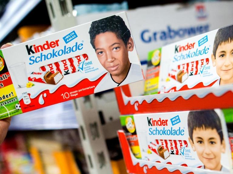 Eine Hand Hält eine Schachtel Kinderschokolade mit dem Kinderbild von Jerome Boateng vor einem Regal im Supermarkt in der Hand.