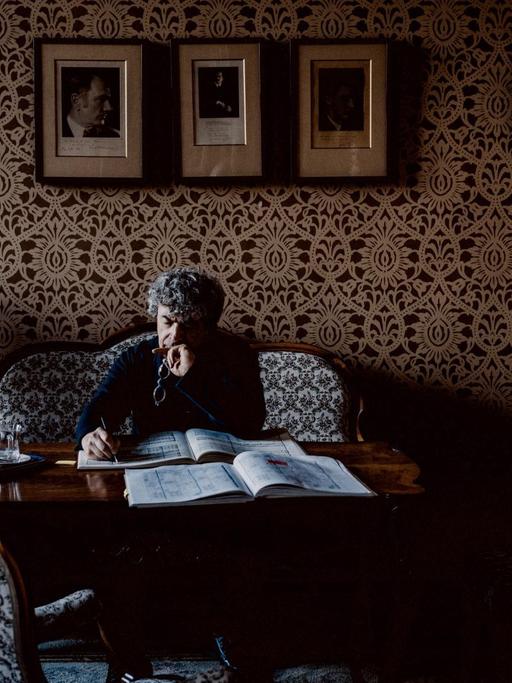 Semyon Bychkov sitzt auf einer antiken Couch, vor sich ein Tisch mit einer Partitur, in die er Notizen schreibt