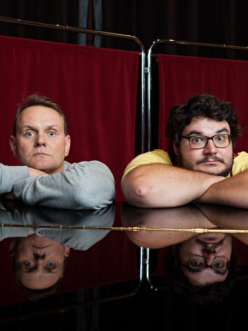 Mit verschränkten Armen sitzen Devid Striesow und Axel Ranisch an einem Klavier.