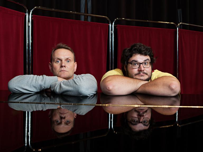Mit verschränkten Armen sitzen Devid Striesow und Axel Ranisch an einem Klavier.