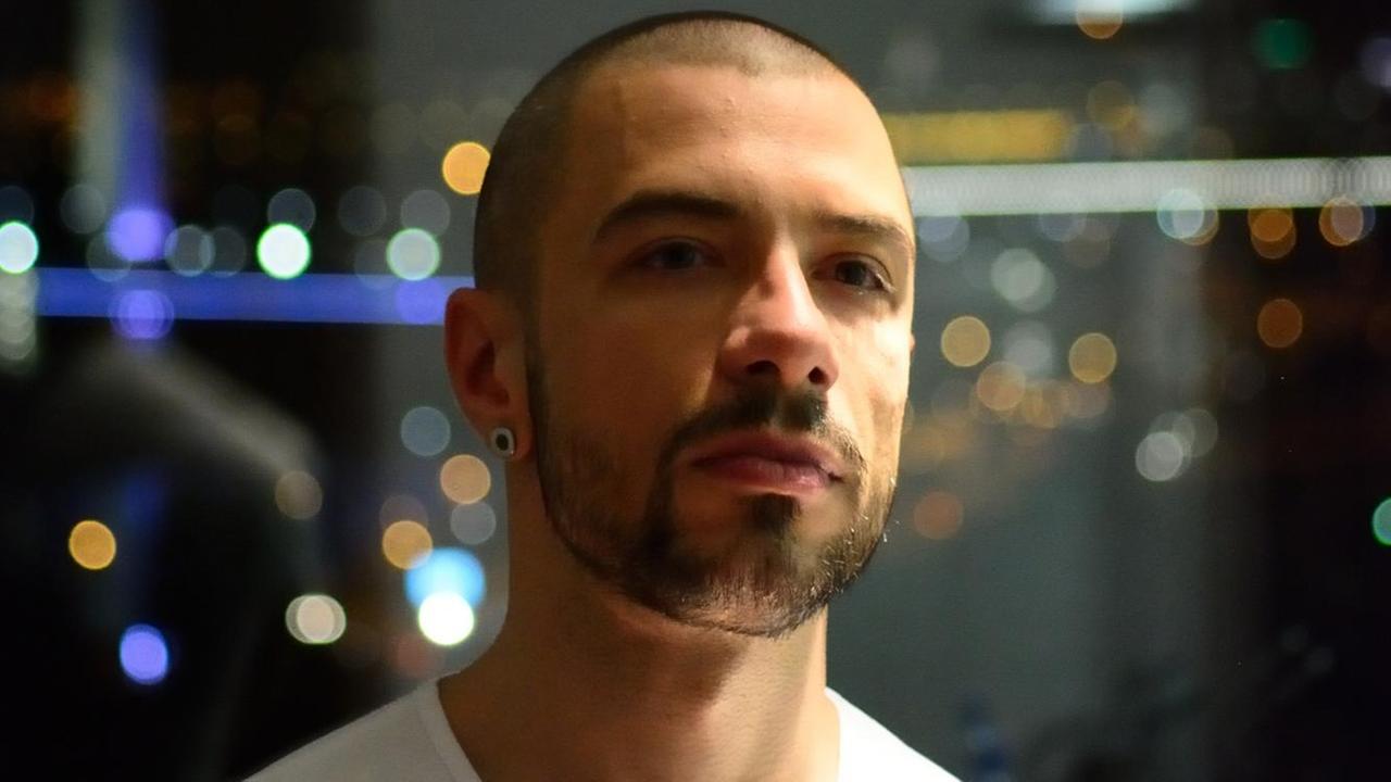 Ein Mann mit weißem T-Shirt ist vor einem Fenster hinter dem ein Nachtpanorama zu sehen ist Ein Mann mit weißem T-Shirt ist vor einem Fenster hinter dem ein Nachtpanorama zu sehen ist