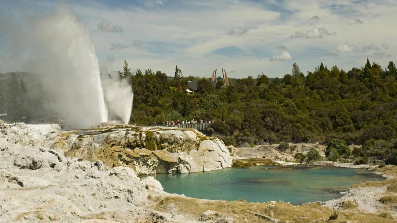 Pohutu-Geysir bei Rotorua in Neuseeland: Eine Waldlandschaft mit einem zischen Geysir bei einem See. In der Nähe halten sich Touristen auf. Pohutu-Geysir bei Rotorua in Neuseeland: Eine Waldlandschaft mit einem zischen Geysir bei einem See. In der Nähe halten sich Touristen auf.