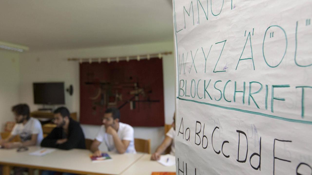 Im Vordergrund ist ein Tafel mit dem Alphabet zu sehen, im Hintergrund sitzen Menschen an Tischen. Im Vordergrund ist ein Tafel mit dem Alphabet zu sehen, im Hintergrund sitzen Menschen an Tischen.
